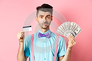 Handsome man in bow-tie showing plastic credit card and money in dollars, making an offer, standing over pink background