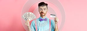 Handsome man in bow-tie showing plastic credit card and money in dollars, making an offer, standing over pink background