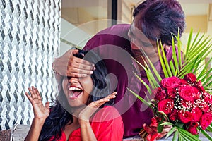 Handsome man closing eyes of his girlfriend to make romantic surprise in restaurant summer tropical cafe urban beach