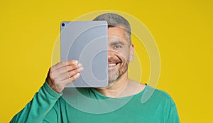 Handsome man cover half of his face hold digital tablet in hand smiling looking at camera isolated on yellow background