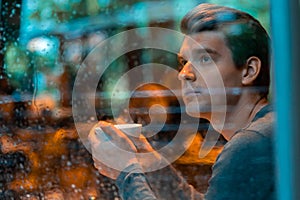 Handsome man drinking tea or coffee on rainy day and looking through the window