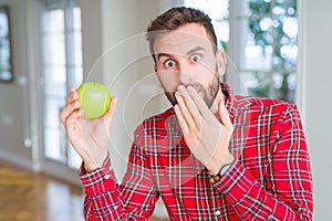 Handsome man eating fresh healthy green apple cover mouth with hand shocked with shame for mistake, expression of fear, scared in
