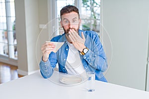 Handsome man eating healthy sandwich cover mouth with hand shocked with shame for mistake, expression of fear, scared in silence,