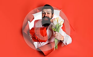 Handsome man with gift box and bouquet of flowers. Businessman with gift and tulips looking through paper hole. Bearded