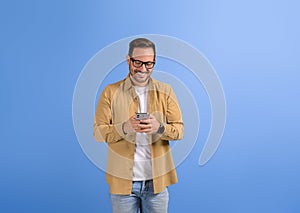 Handsome man in glasses smiling and checking social media apps on smart phone over blue background