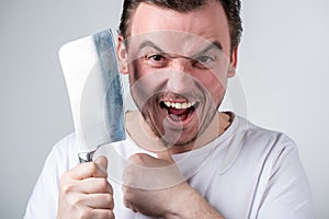 Handsome man grimaces and wants to shave stubble with a kitchen cleaver on a gray background