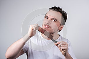Handsome man grimaces and wants to shave stubble with a kitchen cleaver on a gray background