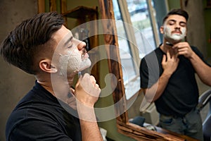 Handsome man hairdresser shaving while looking at mirror