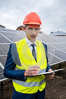 handsome man in a helmet and vest in his hands holding a tablet with records standing near the panels.