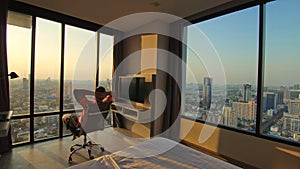 Handsome man in his room in luxury hotel is looking beautiful sunrise. Man in an apartment against background of big