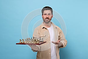 Handsome man holding chessboard with game pieces on light blue background