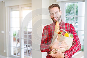 Handsome man holding paper bag full of fresh groceries at home
