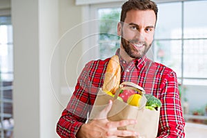 Handsome man holding paper bag full of fresh groceries at home