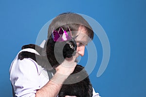 Handsome man holding on sholder black cat in pink shiny crown on blue background with copy space photo