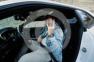 Handsome man in jeans jacket and cap sit at his white muscle car and show Rock and Roll hand sign