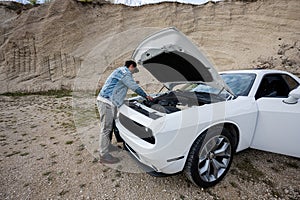 Handsome man in jeans jacket and cap is standing near his white muscle car with opening hood, check oil level of engine