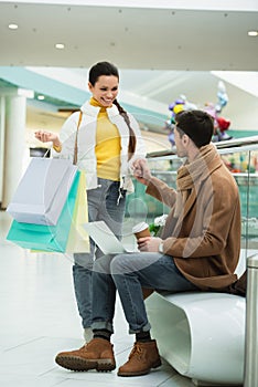 Handsome man with laptop and disposable cup sitting on bench and holding hand of girl with girl with shopping bags