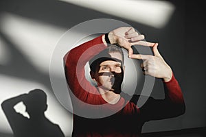 Handsome man looking through a frame formed by his hands in a red sweater on a white background with shadows from the light. Copy