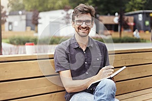 Handsome man making notes in a book sitting outside. Smiling face wearing glasses alone working. Concept of education students