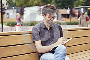 Handsome man making notes in a book sitting outside. Smiling face wearing glasses alone working. Concept of education students