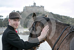 Handsome man, Male Horse Rider pets his horse on beach, wearing traditional flat cap, white trousers, red polo shirt