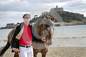 Handsome man, Male Horse Rider walking with his horse on beach, wearing traditional flat cap, white trousers, red polo shirt