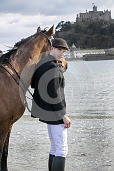 Handsome man, Male Horse Rider walking with his horse on beach, wearing traditional flat cap, white trousers, red polo shirt