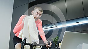 Handsome man with mustache doing fitness cardio workout exercises on smart cycling trainer and wiping face with towel. Male athlet