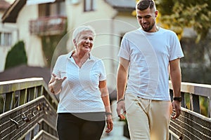 A handsome man and an older woman share a serene walk in nature, crossing a beautiful bridge against the backdrop of a