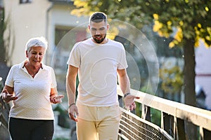 A handsome man and an older woman share a serene walk in nature, crossing a beautiful bridge against the backdrop of a