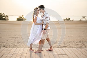 Handsome man in a plaid shirt embracing his beautiful pregnant wife in a white dress at beach sunset.