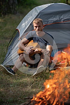 Handsome man playing guitar in forest with bonfire. Romantic guy camping outdoors and sitting near tent. Man with guitar