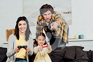Handsome man pointing with finger near while cheerful wife and kid playing video game at home.