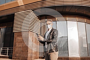 Handsome man in a protective mask on his face with glasses with a smartphone on the street of a big city. Businessman talking on