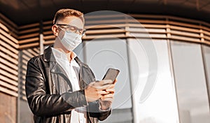 Handsome man in a protective mask on his face with glasses with a smartphone on the street of a big city. Businessman talking on