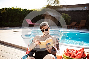 Handsome man reading book, lying on chaise near swimming pool.