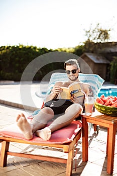 Handsome man reading book, lying on chaise near swimming pool.