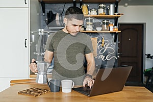 Handsome man remotely works with a laptop and pours coffee in the interior of a home kitchen in the morning.