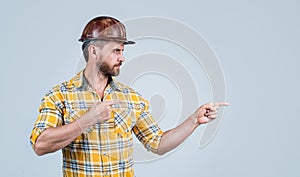 handsome man repairman in construction safety helmet and checkered shirt on building site pointing finegr, copy space