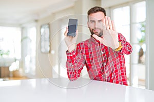 Handsome man showing smartphone screen with open hand doing stop sign with serious and confident expression, defense gesture