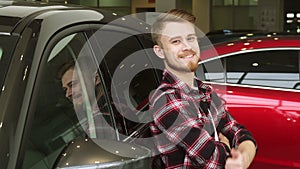 Handsome man showing thumbs up leaning on a new car at the dealership