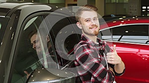 Handsome man showing thumbs up leaning on a new car at the dealership