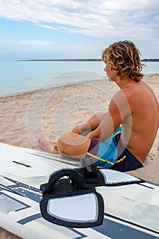 Handsome man sits on the beach with white blank surfing board wait for wave to surf spot at sea ocean shore. Concept of