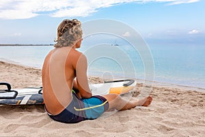 Handsome man sits on the beach with white blank surfing board wait for wave to surf spot at sea ocean shore. Concept of