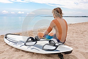 Handsome man sits on the beach with white blank surfing board wait for wave to surf spot at sea ocean shore. Concept of