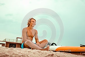 Handsome man sits on the beach with white blank surfing board wait for wave to surf spot at sea ocean shore. Concept of