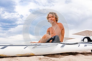 Handsome man sits on the beach with white blank surfing board wait for wave to surf spot at sea ocean shore. Concept of