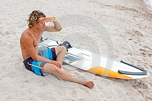 Handsome man sits on the beach with white blank surfing board wait for wave to surf spot at sea ocean shore. Concept of