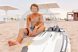 Handsome man sits on the beach with white blank surfing board wait for wave to surf spot at sea ocean shore. Concept of