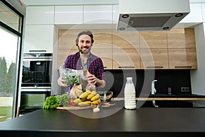 Handsome man smiles looking at camera, holding greens while unpacking eco paper bag with healthy food, standing at kitchen